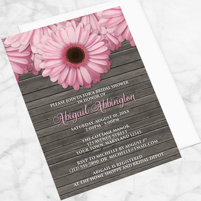 Rustic Pink Daisy Brown Wood Bridal Shower Invitation shown at an angle on a marble surface with a white envelope.