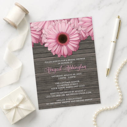 Rustic Pink Daisy Brown Wood Bridal Shower Invitation shown on a marble surface with decorative items.