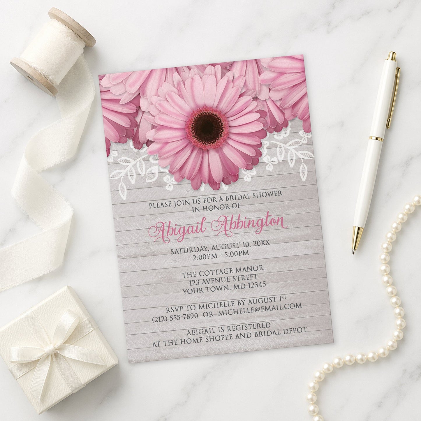 Rustic Pink Daisy Gray Wood Bridal Shower Invitation on a marble surface with a white gift box, pen, ribbon, and pearls.