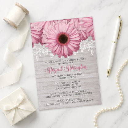 Rustic Pink Daisy Gray Wood Bridal Shower Invitation on a marble surface with a white gift box, pen, ribbon, and pearls.
