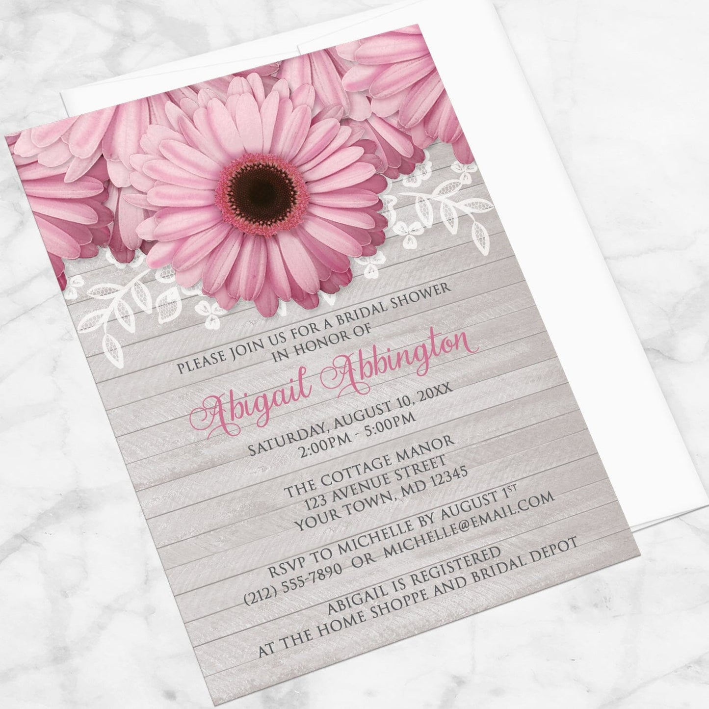 Bridal shower invitation with pink daisies, white lace, and soft gray wood, displayed at an angle with a white envelope on a marble surface