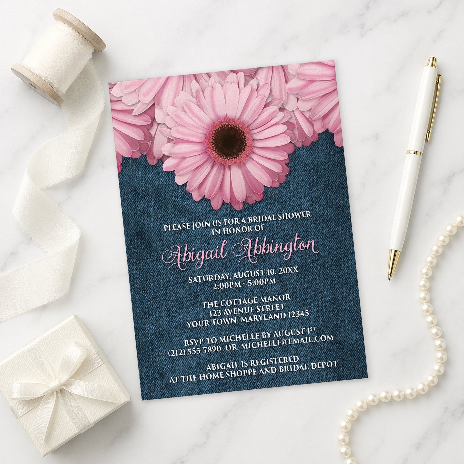 Rustic Pink Daisy and Denim Bridal Shower Invitation shown on a marble surface with a white gift box, pen, ribbon, and pearls.
