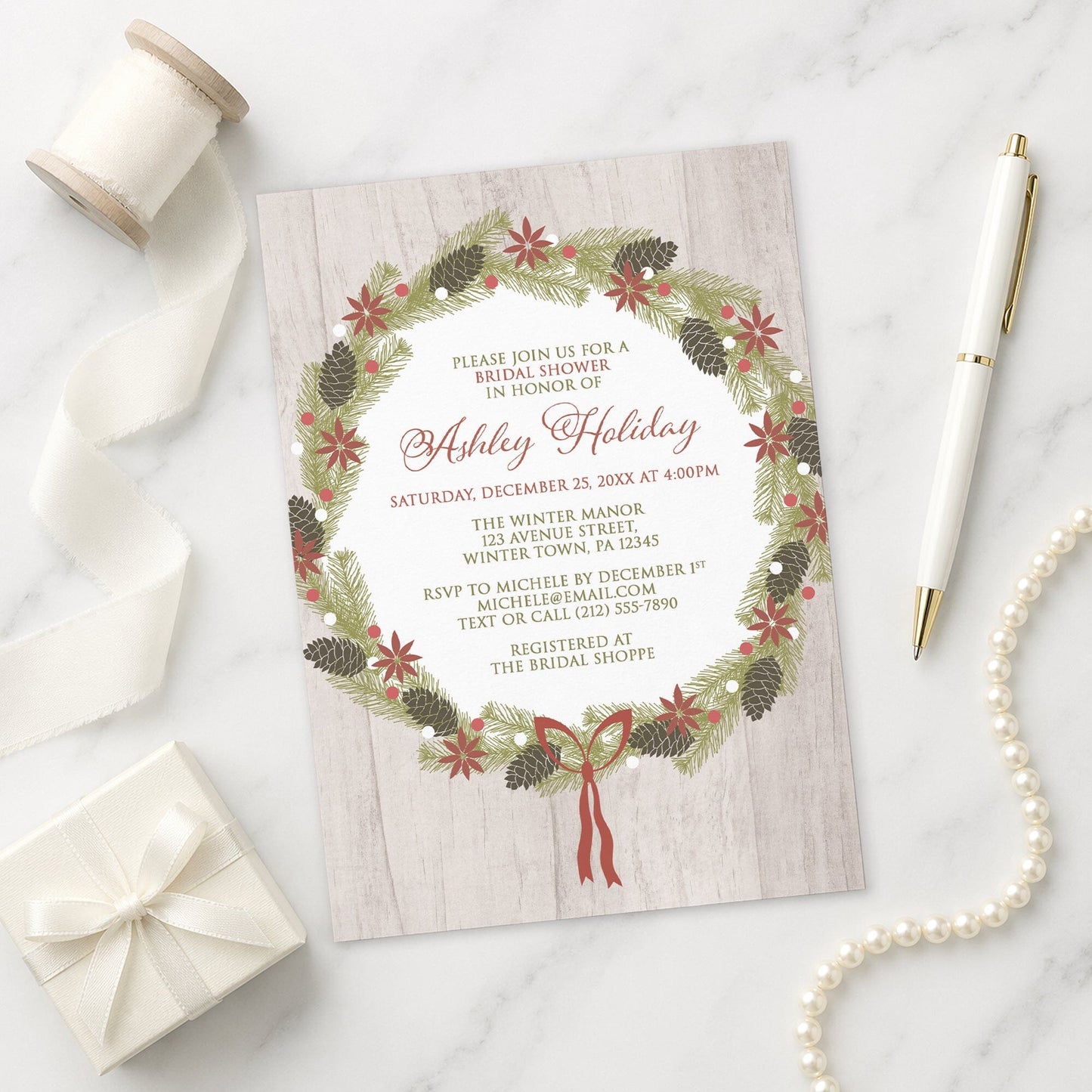Rustic Poinsettia Pine Cone Wreath Bridal Shower Invitation on a marble surface with a white gift box, pen, ribbon, and pearls.