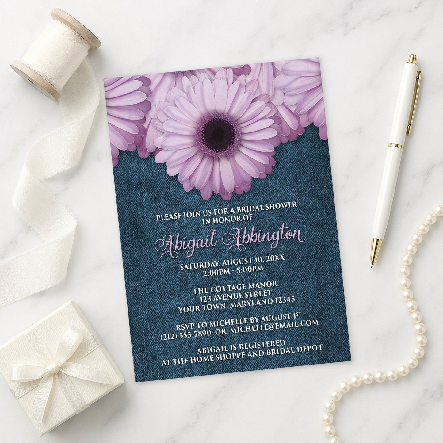 Rustic Purple Daisy Denim Bridal Shower Invitation shown on a marble surface with a white gift box, pen, ribbon, and pearls.