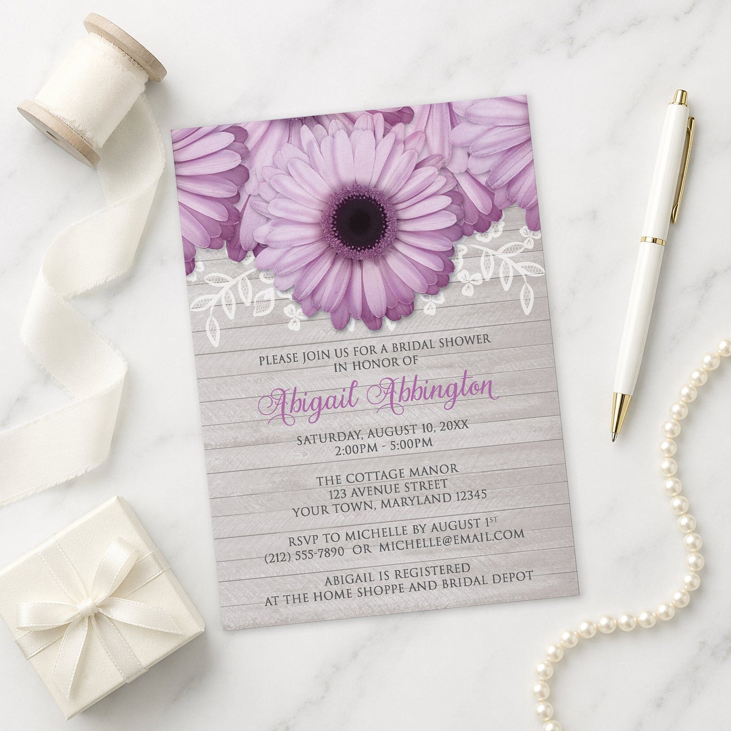 Rustic Purple Daisy Gray Wood Bridal Shower Invitation on a marble surface with a white gift box, pen, ribbon, and pearls.