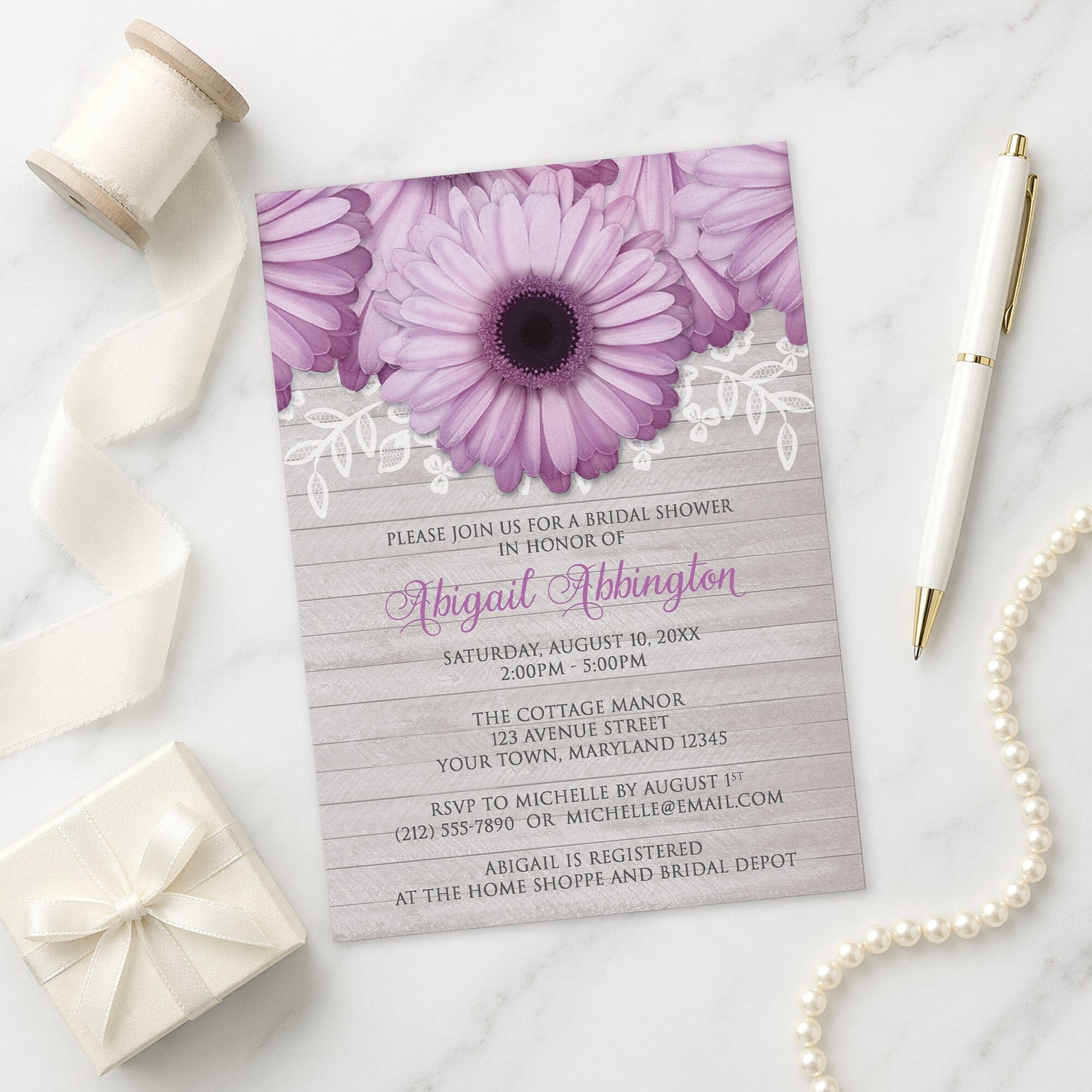 Rustic Purple Daisy Gray Wood Bridal Shower Invitation on a marble surface with a white gift box, pen, ribbon, and pearls.