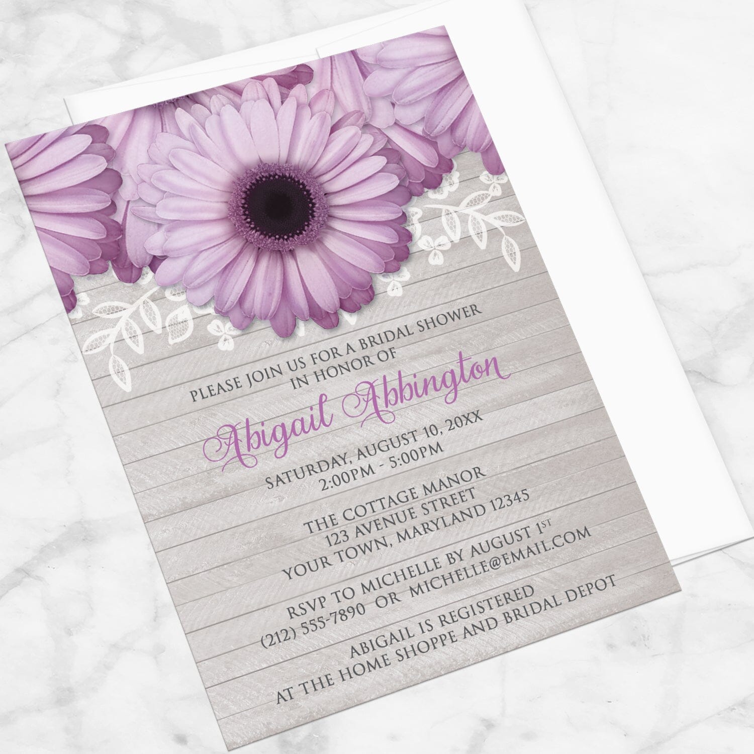 Bridal shower invitation with purple daisies, lace, and gray wood, displayed at an angle with a white envelope on a marble surface