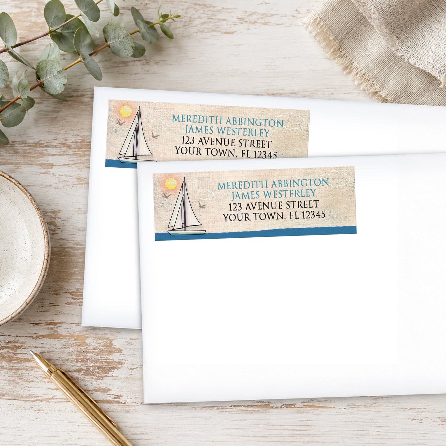 Nautical-themed sailboat address labels on envelopes with a pen and eucalyptus branch on a wooden surface.