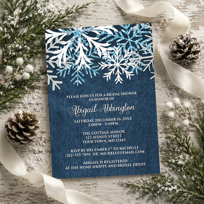 Rustic Snowflake Denim Winter Bridal Shower Invitation shown on a painted wood background with winter greenery, pine cones, and ribbons.