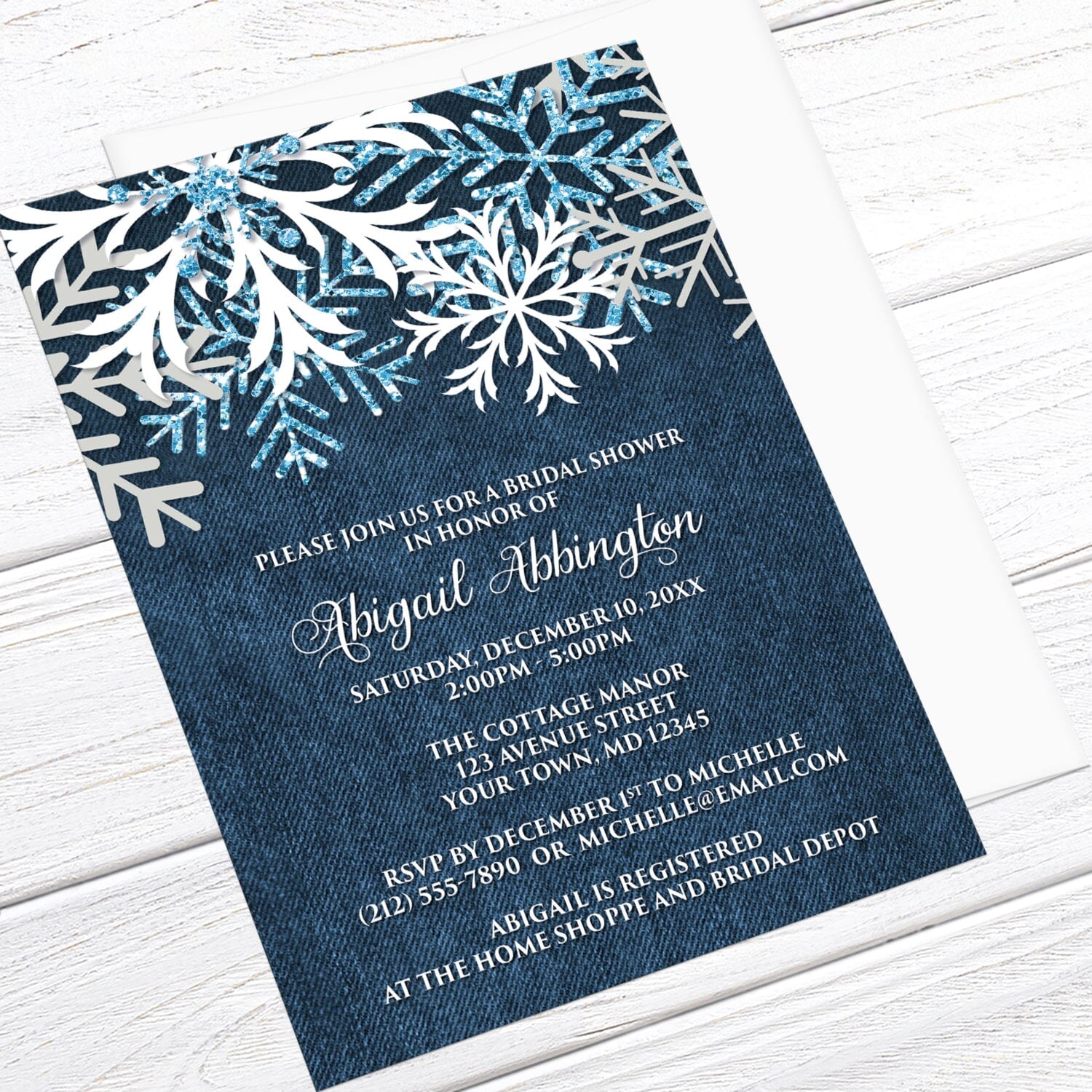 Rustic Snowflake Denim Winter Bridal Shower Invitation shown at an angle in a light wood surface with a white envelope.