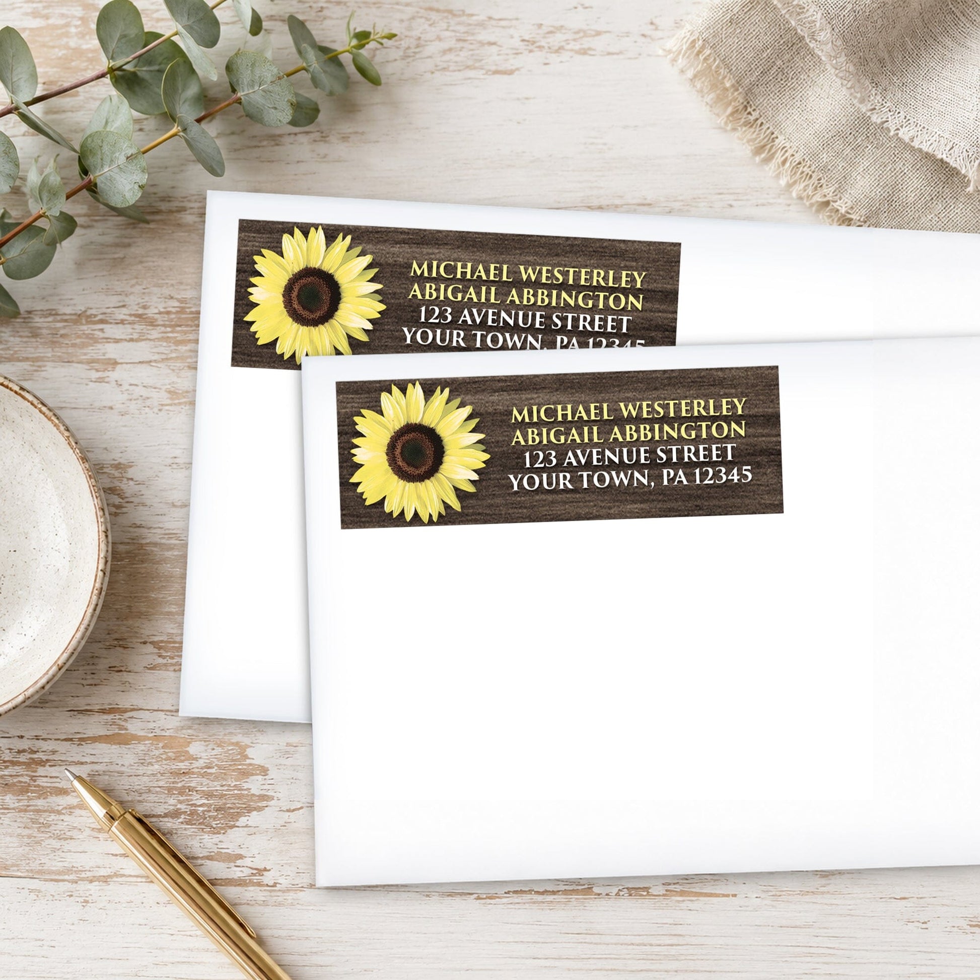 Address labels with a sunflower design with rustic brown wood, on white envelopes, shown on a wooden surface with eucalyptus leaves and a pen.