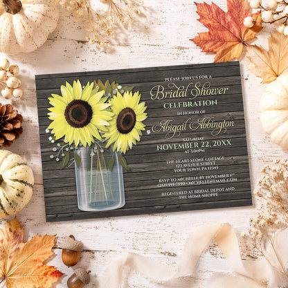 A styled flat-lay of a bridal shower invitation card featuring a rustic dark wood background, yellow sunflowers in a glass mason jar, and light yellow, green, and white text. The card is surrounded by autumn-themed props including pumpkins, fall leaves, acorns, and dried flowers on a whitewashed wooden surface.