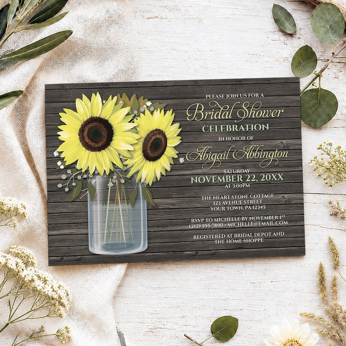 A rustic bridal shower invitation card styled in a flat-lay arrangement on a white wood surface with linen fabric, greenery, and small white flowers. The invitation features a dark wood plank background, two large yellow sunflowers in a clear glass mason jar, and light yellow, green, and white text.