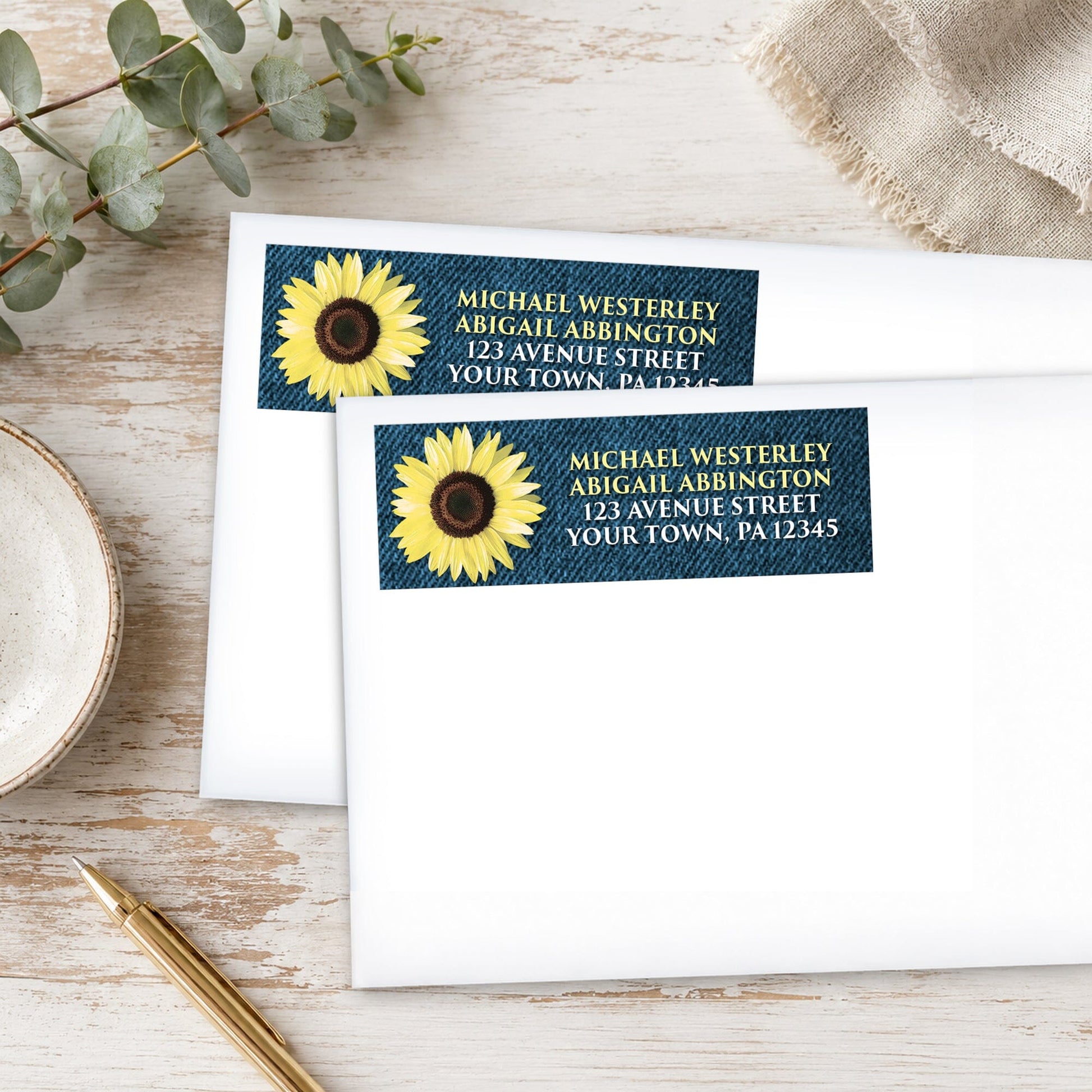 Address labels with sunflower and denim design on white envelopes, placed on a wooden surface with eucalyptus leaves and a pen.