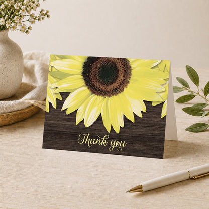 Rustic Sunflower and Wood Thank You Card shown on a tabletop with a vase, plants, and a pen.