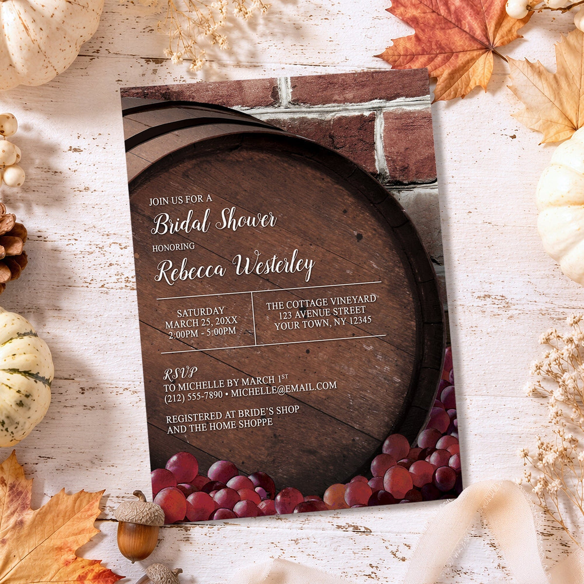 A styled bridal shower invitation featuring a rustic wine barrel and red grapes design, laid flat on a whitewashed wooden surface surrounded by autumn leaves, pumpkins, and acorns.