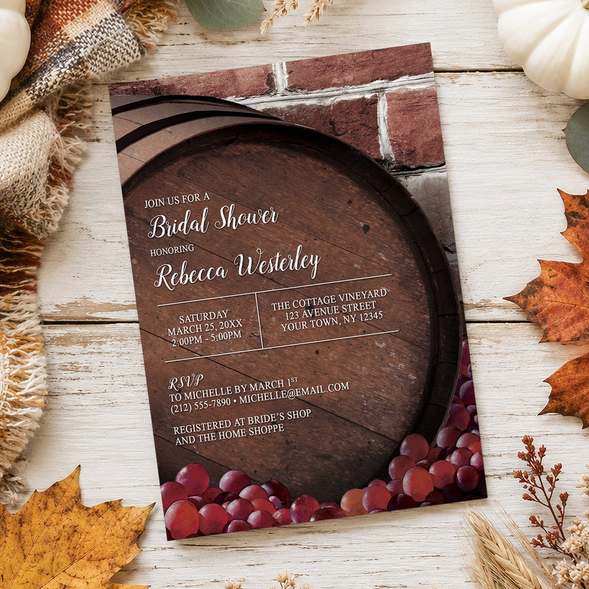 A rustic bridal shower invitation styled on a white wooden surface with autumn leaves, burlap, pumpkins, and a blanket. The invitation features a dark wood barrel design and a brick wall background, with red grapes at the bottom and white text over the barrel.