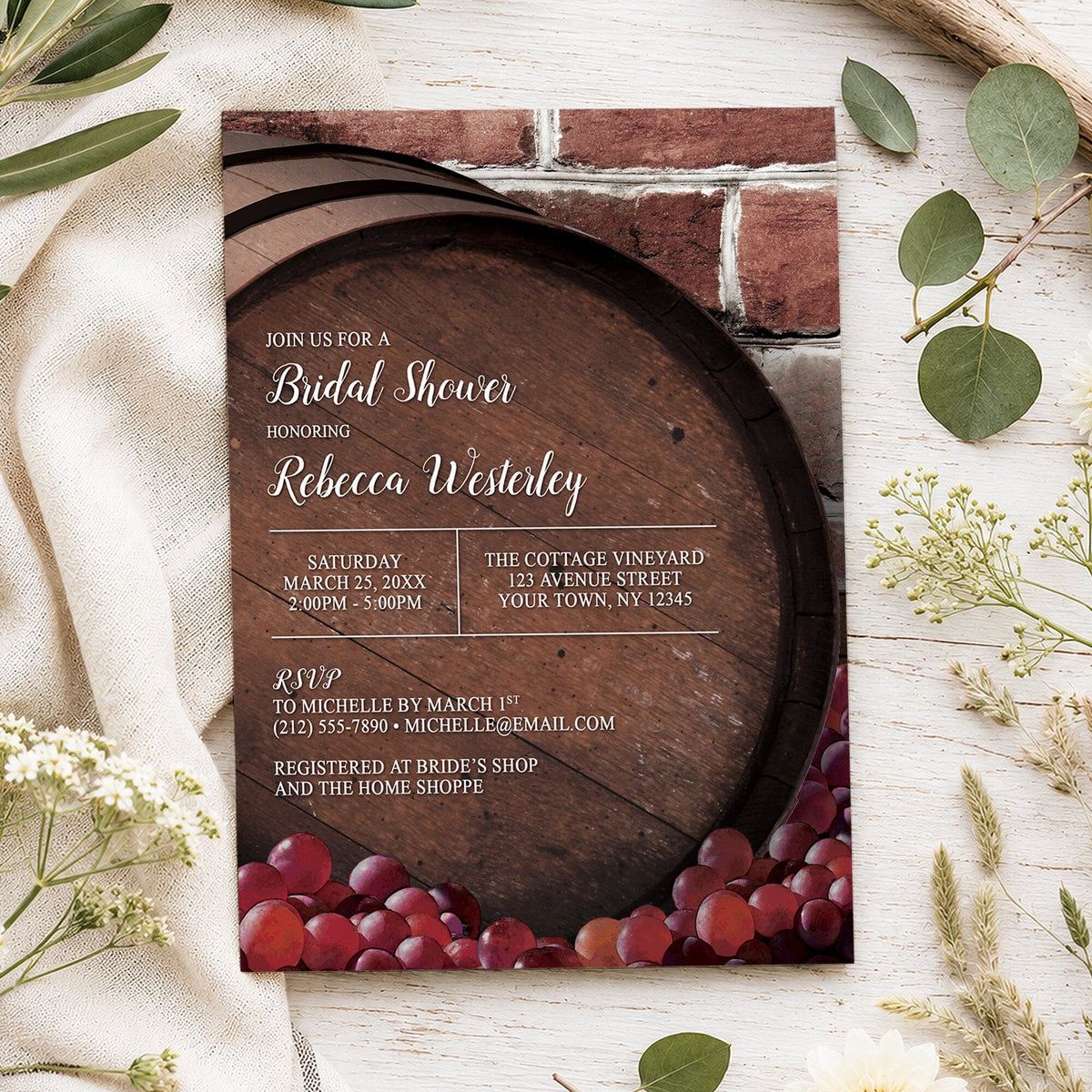 A rectangular bridal shower invitation styled in a flat lay on a white fabric surface with scattered green leaves and small white flowers. The card features a rustic wine barrel and brick background with red grapes along the bottom and white event text in white over the barrel.