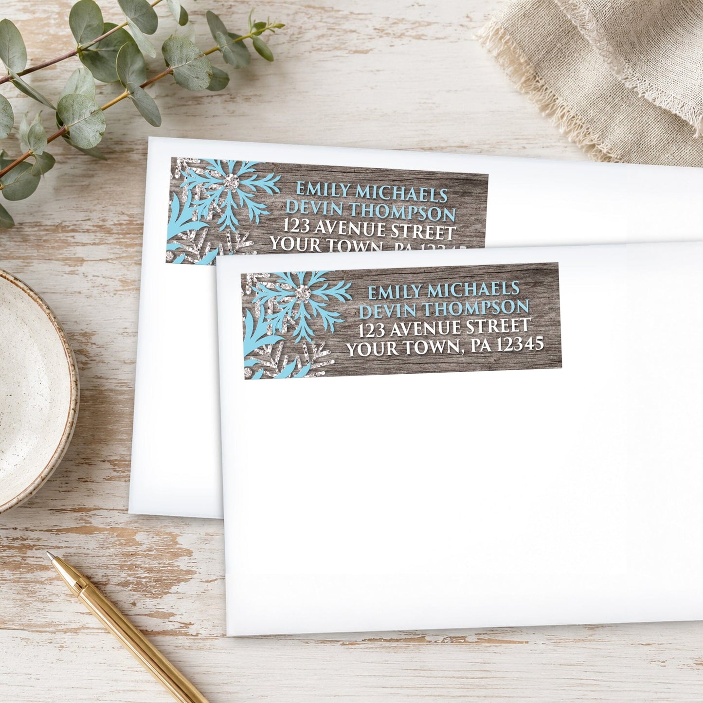 Rustic Winter Wood Blue Snowflake Address Labels, shown on white envelopes, on a wooden surface with eucalyptus leaves and a pen.