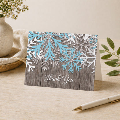 Rustic Winter Wood Blue Snowflake Thank You Card shown on a desk with a vase, plants, and a pen.