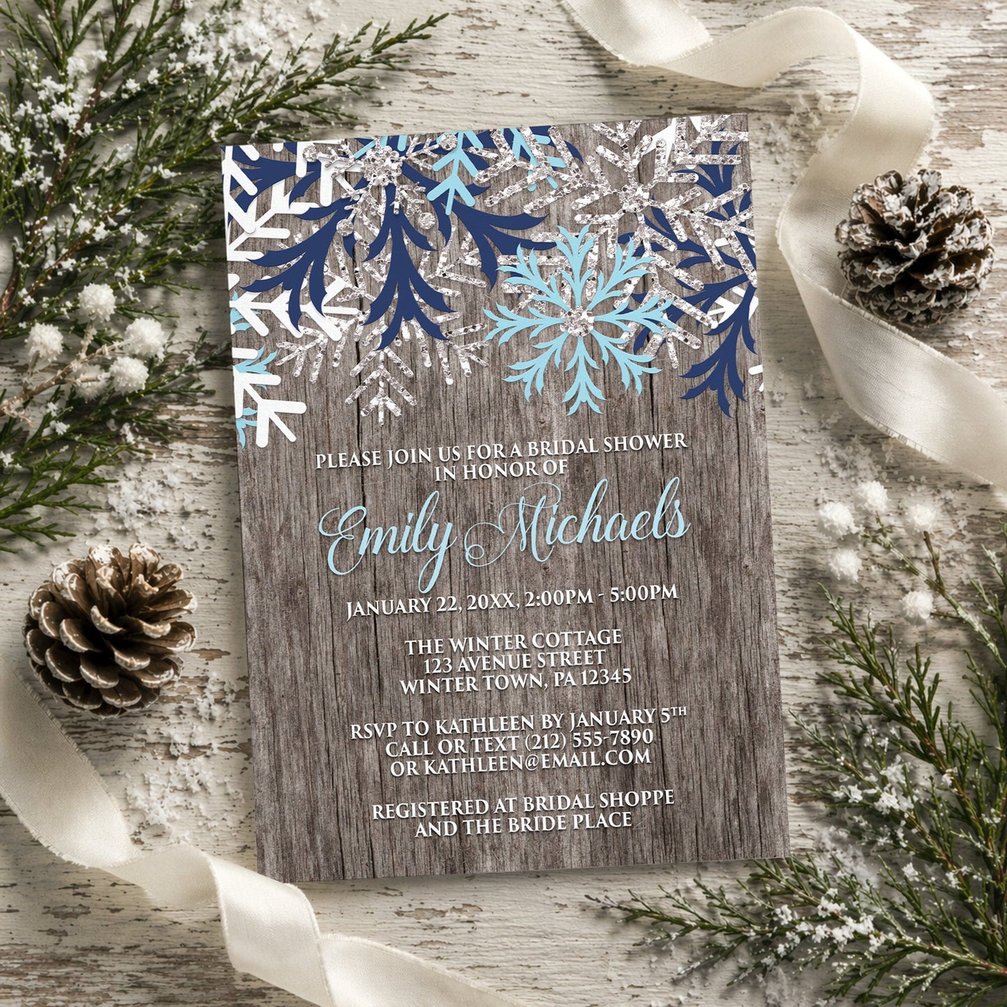 Rustic Winter Wood Navy Aqua Snowflake Bridal Shower Invitation shown on a light painted wooden surface with winter greenery, pine cones, and ribbons.