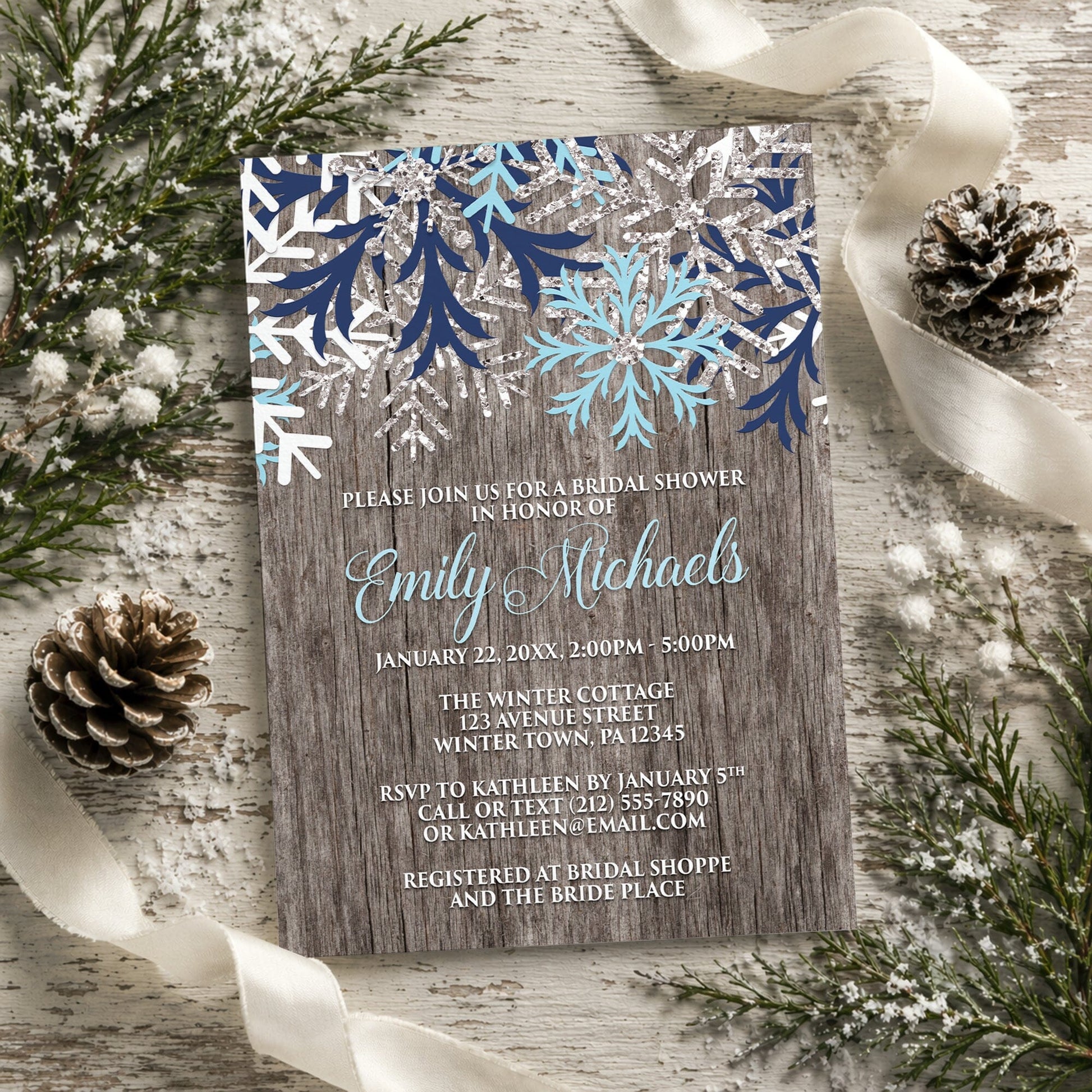 Rustic Winter Wood Navy Aqua Snowflake Bridal Shower Invitation shown on a light painted wooden surface with winter greenery, pine cones, and ribbons.