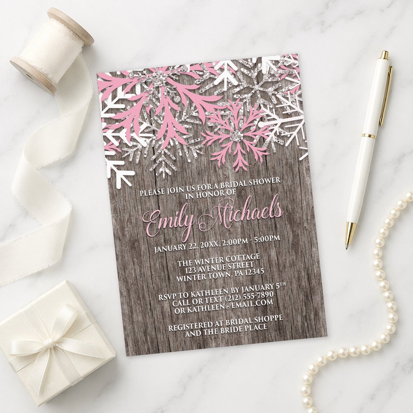 Rustic Winter Wood Pink Snowflake Bridal Shower Invitation shown on a marble surface with a white gift box, pen, ribbon, and pearls.