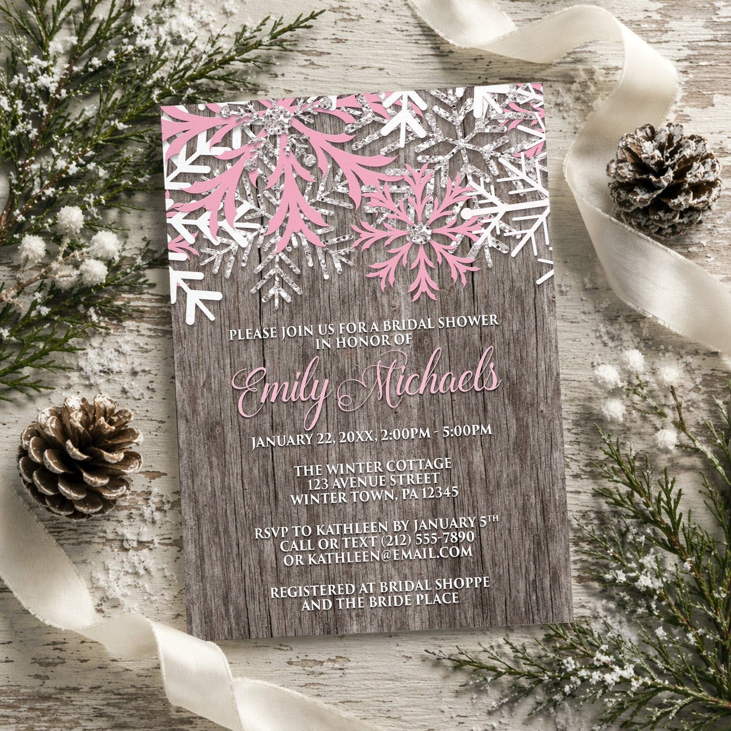 Rustic Winter Wood Pink Snowflake Bridal Shower Invitation shown on a painted wooden surface with winter greenery, pine cones, and ribbons.