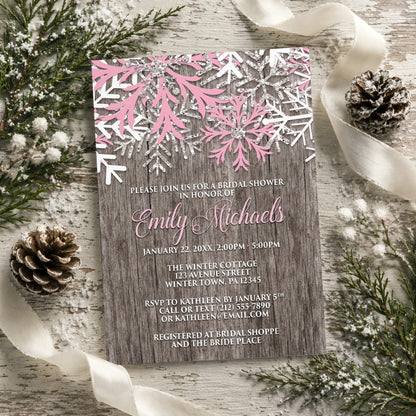 Rustic Winter Wood Pink Snowflake Bridal Shower Invitation shown on a painted wooden surface with winter greenery, pine cones, and ribbons.
