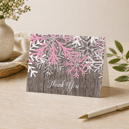 Rustic Winter Wood Pink Snowflake Thank You Card shown on a desk with a vase, plants, and a pen.