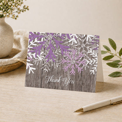 Rustic Winter Wood Purple Snowflake Thank You Card shown on a desk with a vase, plants, and a pen.