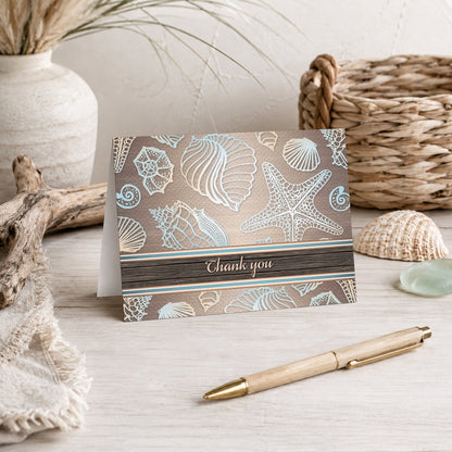 Rustic Wood Beach Seashell Thank You Card shown on a beach house tabletop with coastal decor and a pen.