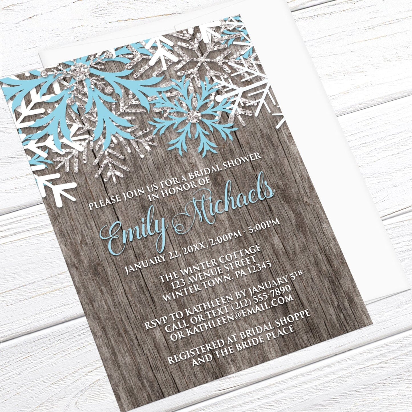 Rustic Winter Wood Blue Snowflake Bridal Shower Invitation shown at an angle on a light wood surface with a white envelope.