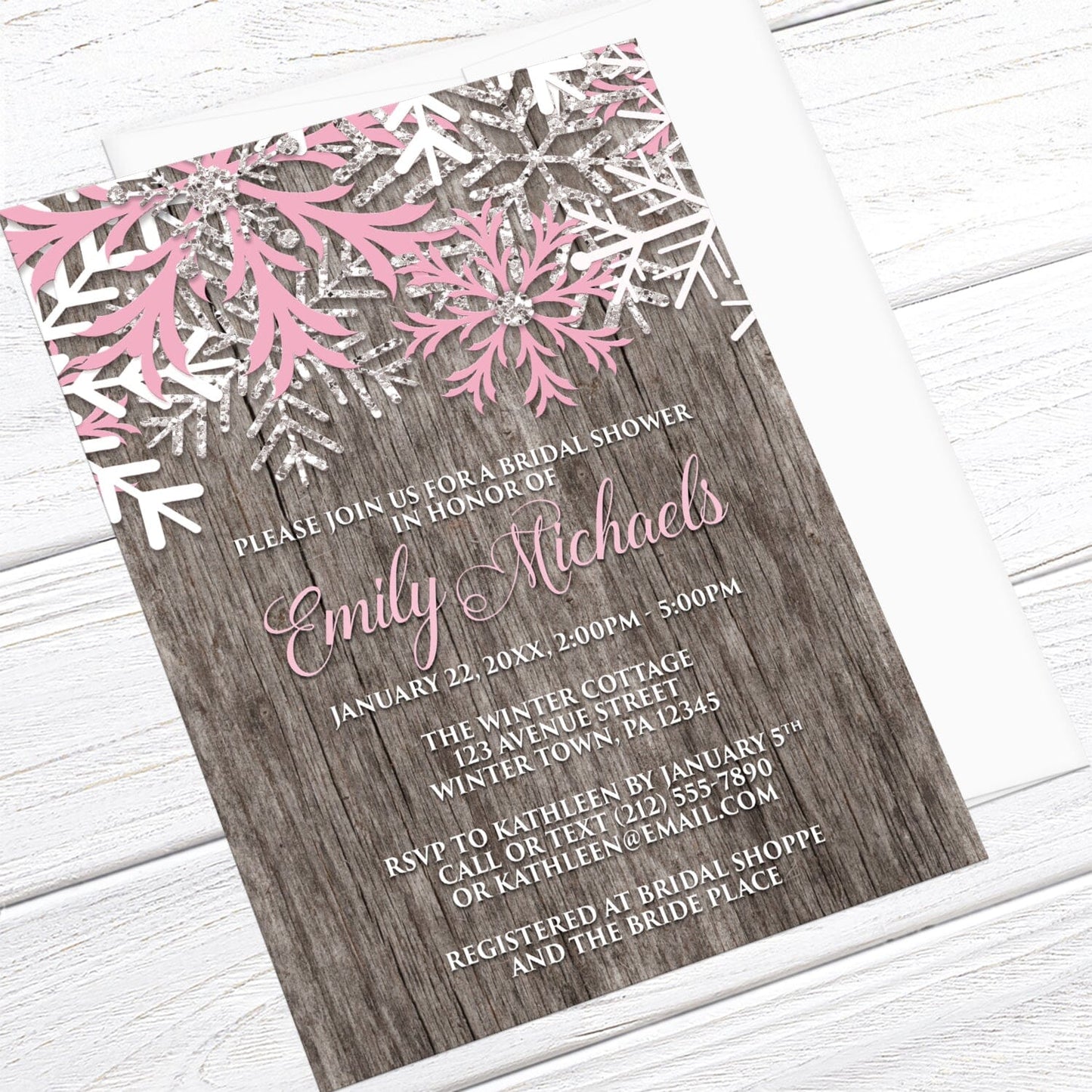 Rustic Winter Wood Pink Snowflake Bridal Shower Invitation shown at an angle on a light wood surface with a white envelope.