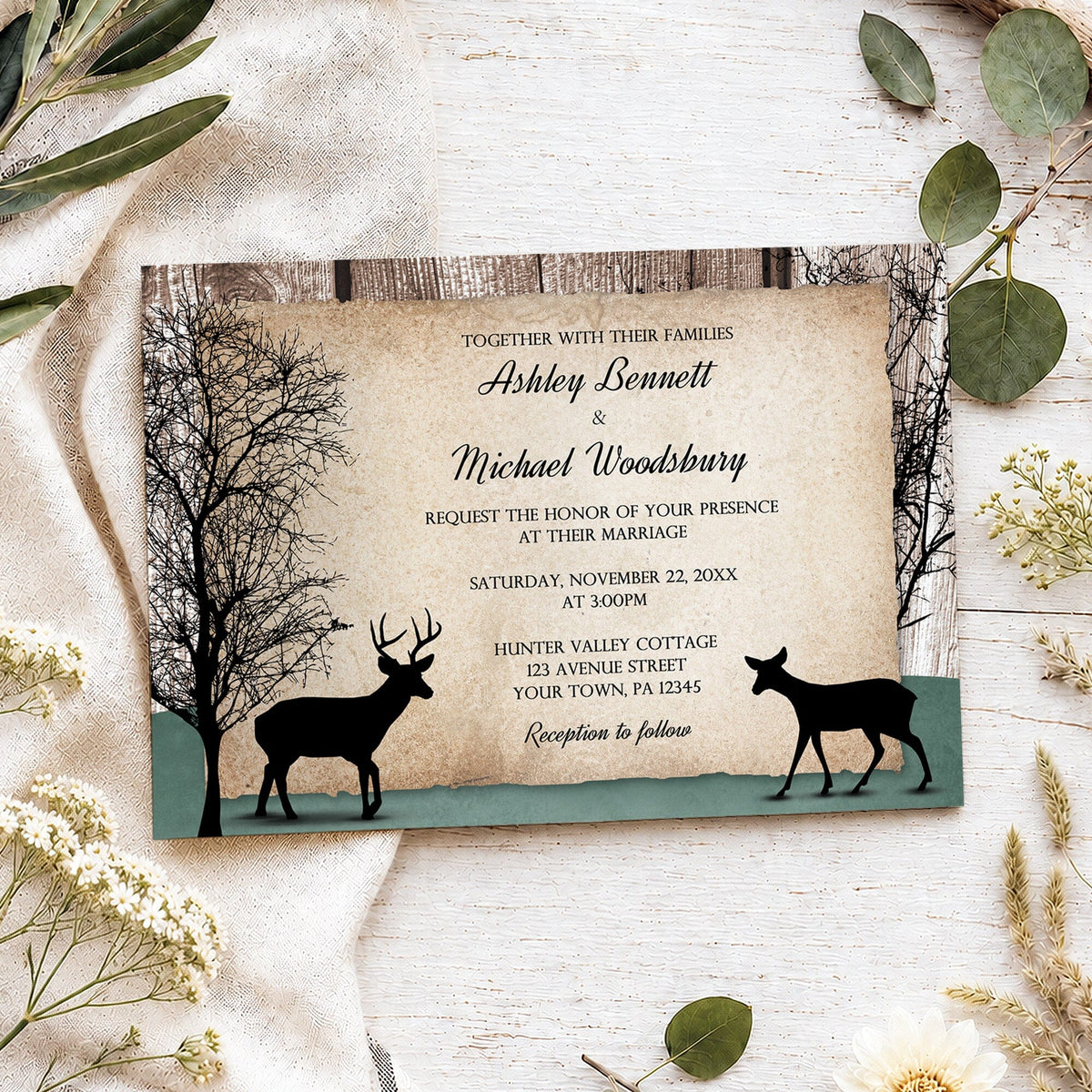A rustic deer wedding invitation styled in a flat-lay on a white wood background with linen fabric and greenery. The invitation features a weathered wood border, a tan parchment-styled center, black silhouettes of trees and deer, and a hunter green ground section.