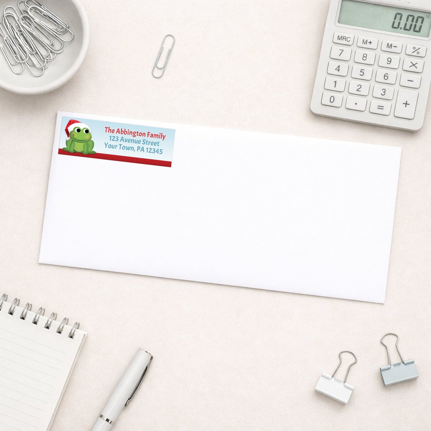 Santa Hat Frog Christmas Address Label shown on a white envelope with office supplies