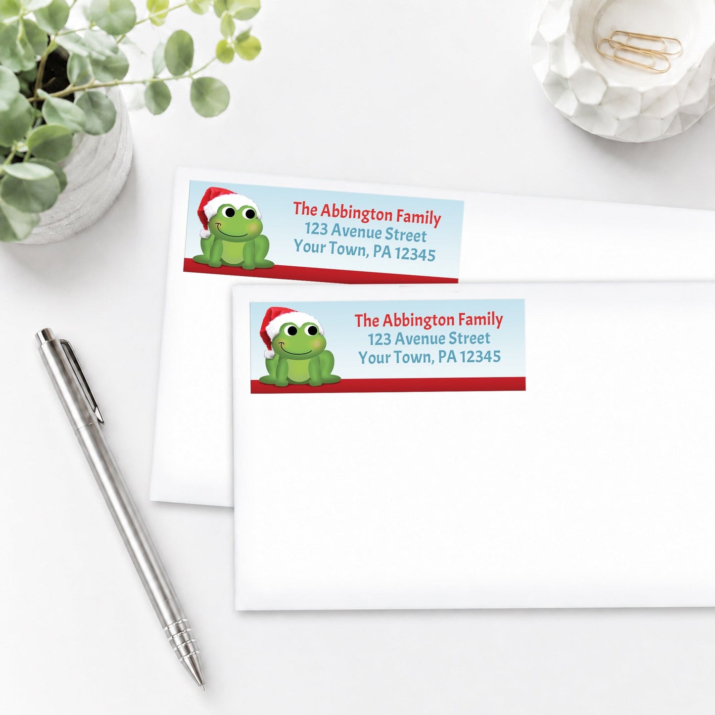 Santa Hat Frog Christmas Address Labels, shown on white envelopes, on a white surface with a plant and a pen.