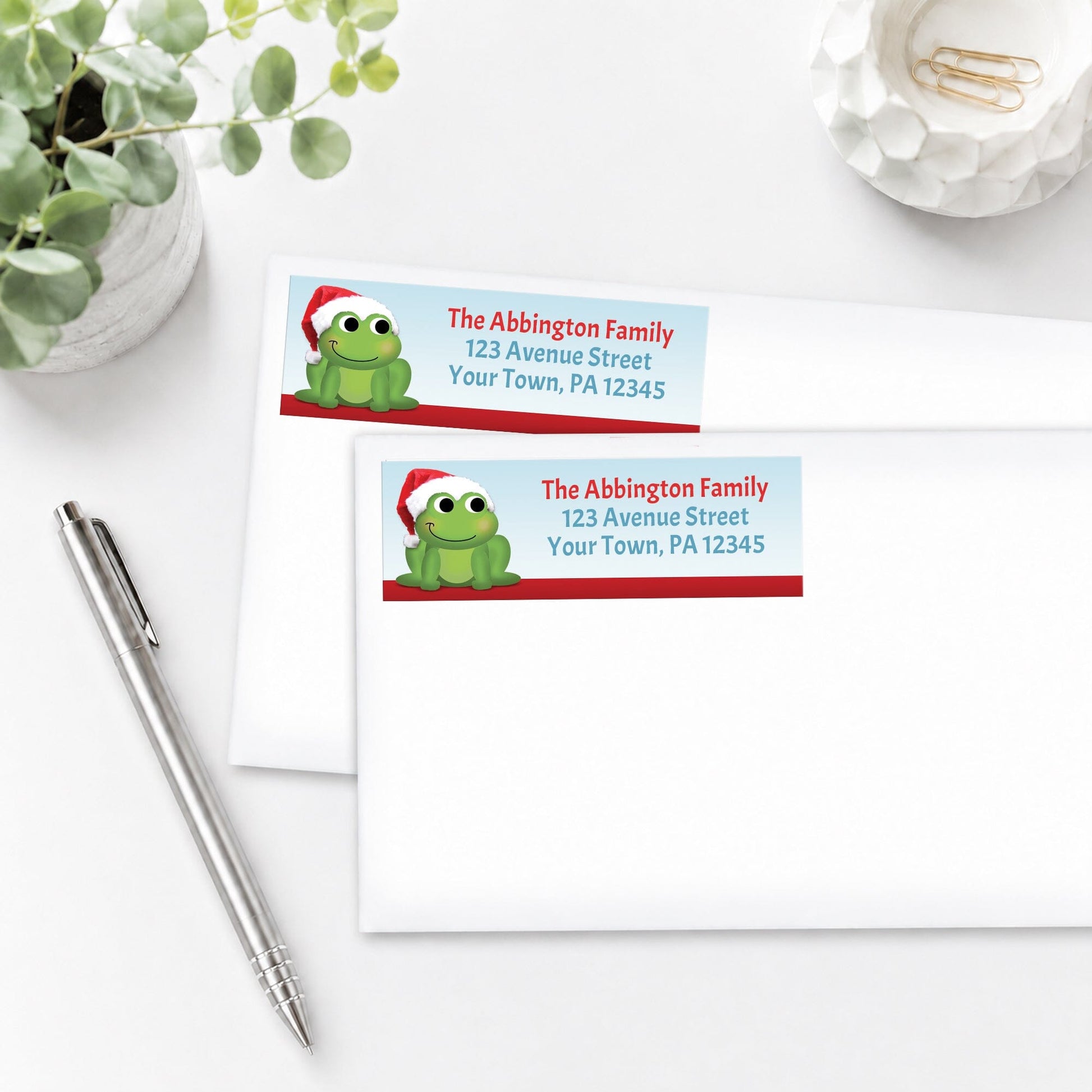 Santa Hat Frog Christmas Address Labels, shown on white envelopes, on a white surface with a plant and a pen.