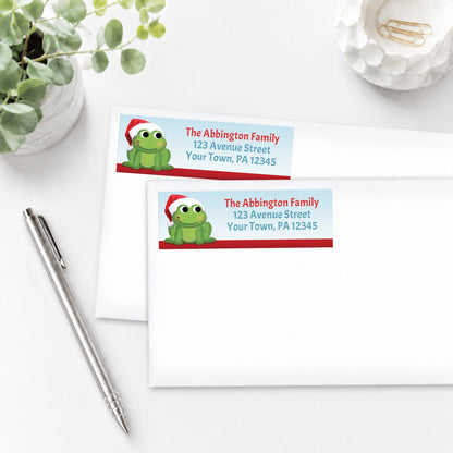 Santa Hat Frog Christmas Address Labels, shown on white envelopes, on a white surface with a plant and a pen.