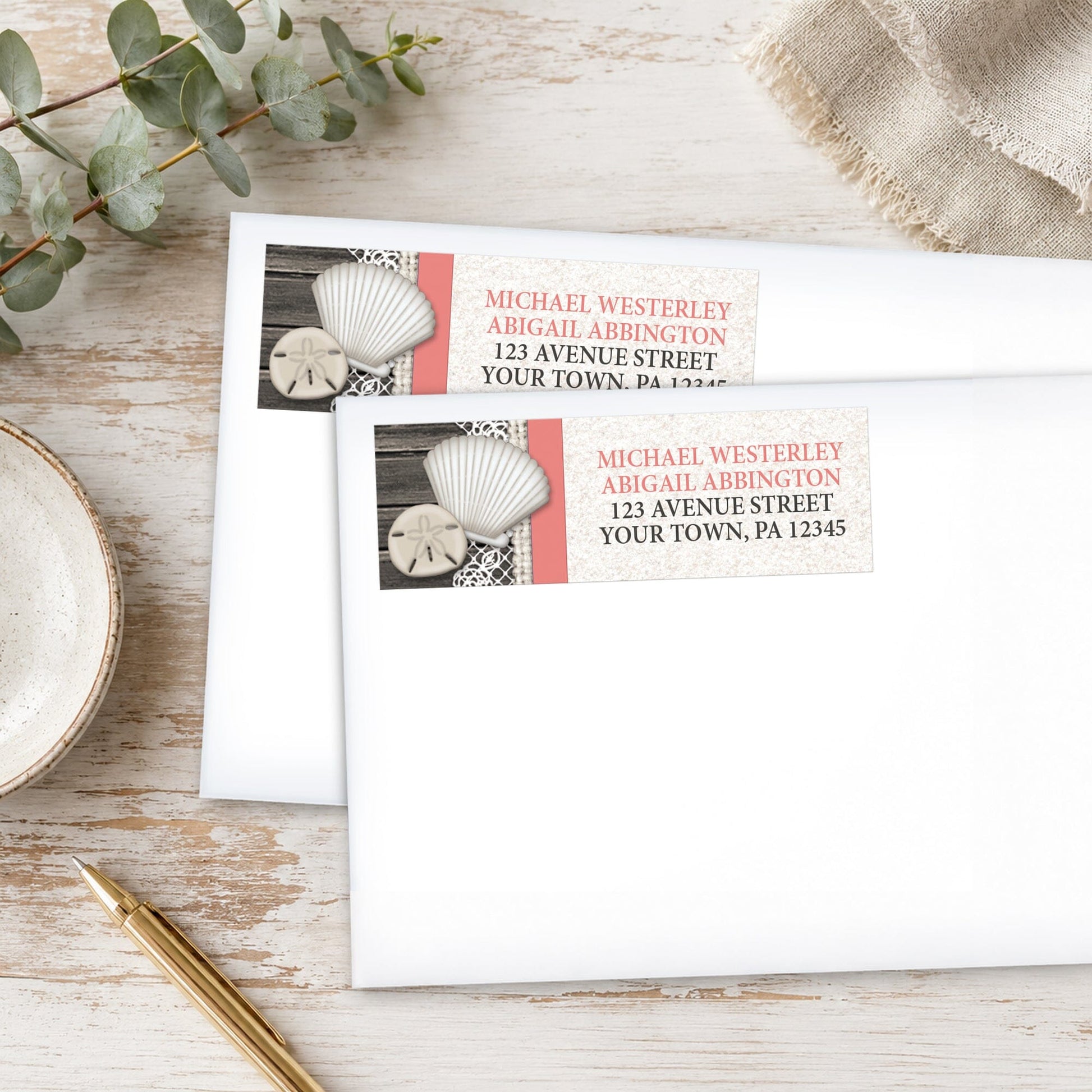 Seashell Lace Wood and Sand Coral Beach Address Labels, shown on white envelopes, on a wooden surface with eucalyptus leaves and a pen.