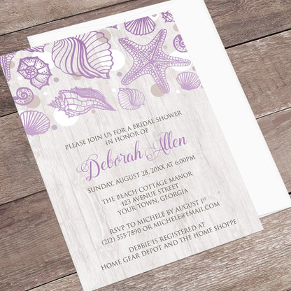 Seashell Whitewashed Wood Purple Beach Bridal Shower Invitation shown at an angle on a wooden surface with a white envelope.