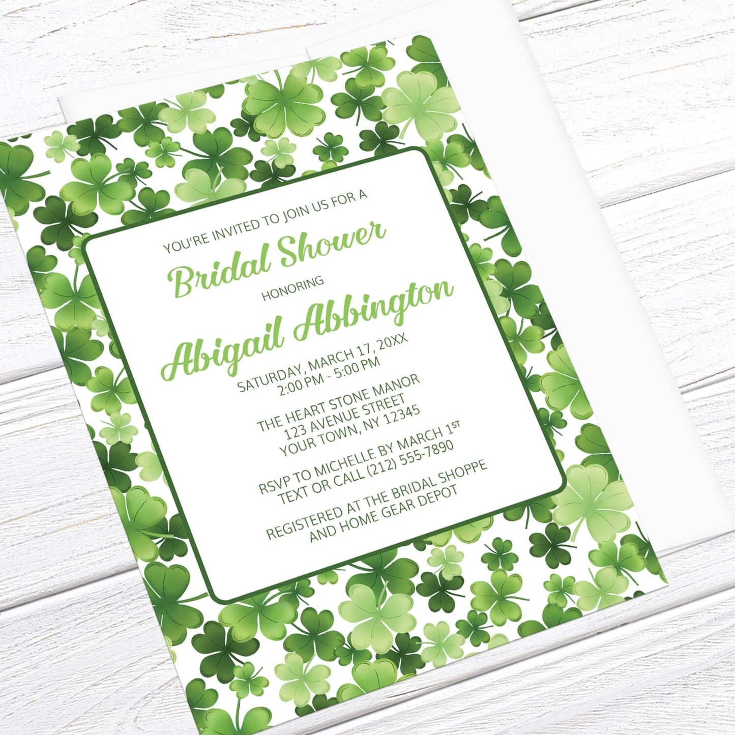Bridal shower invitation with green shamrock pattern, displayed at an angle with a white envelope on a white wood surface