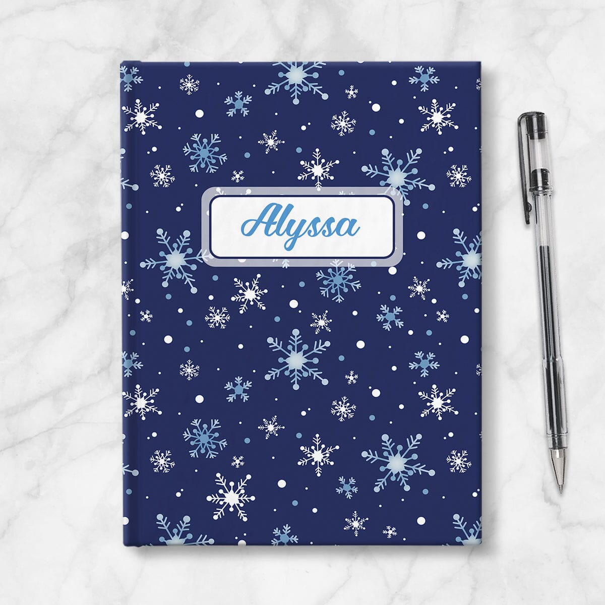 Personalized Snowflakes on Blue Journal at Artistically Invited. A custom book designed with a pattern of whimsically drawn white and blue snowflakes over a navy blue background that wraps around the journal. Your personalized name is custom printed in a blue script font within a white rectangular frame design over the snowflakes pattern. Book is placed next to a pen.