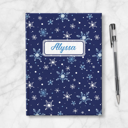 Personalized Snowflakes on Blue Journal at Artistically Invited. A custom book designed with a pattern of whimsically drawn white and blue snowflakes over a navy blue background that wraps around the journal. Your personalized name is custom printed in a blue script font within a white rectangular frame design over the snowflakes pattern. Book is placed next to a pen.
