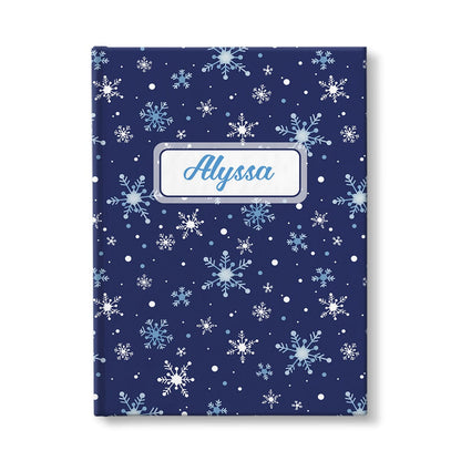 Personalized Snowflakes on Blue Journal at Artistically Invited. A custom book designed with a pattern of whimsically drawn white and blue snowflakes over a navy blue background that wraps around the journal. Your personalized name is custom printed in a blue script font within a white rectangular frame design over the snowflakes pattern.