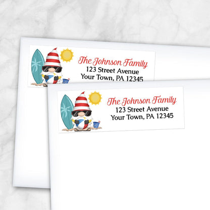 Summer Beach Gnome Address Labels at Artistically Invited. Example of labels on envelopes. These address labels are designed with an illustration of a cute gnome holding a beach ball with a surfboard and a bucket in the sand, and a sun above. Your personalized return address is custom printed in red and black over white to the right of the beach-themed gnome.