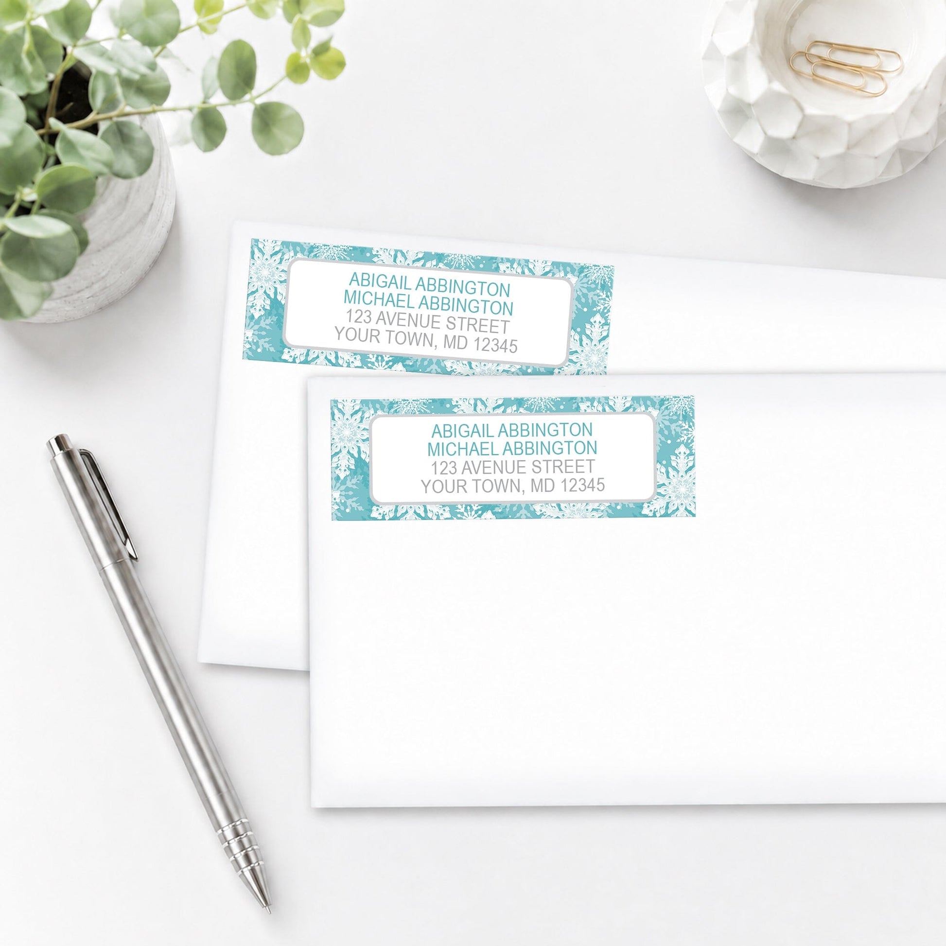 Two personalized Teal Snowflake Winter Address Labels shown on white envelopes, on a white surface with a plant and a pen.