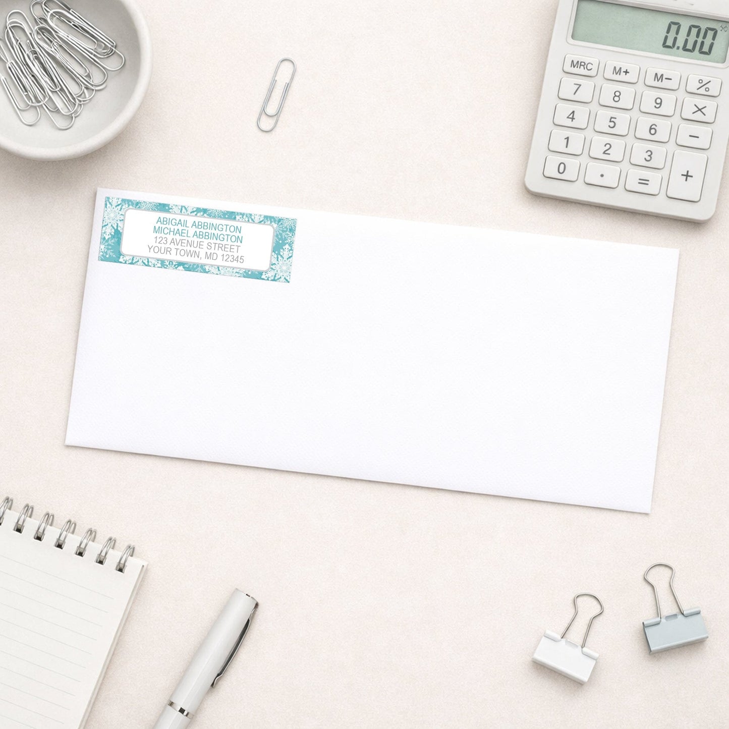 Teal Snowflake Winter Return Address Label shown on a white envelope with everyday office supplies