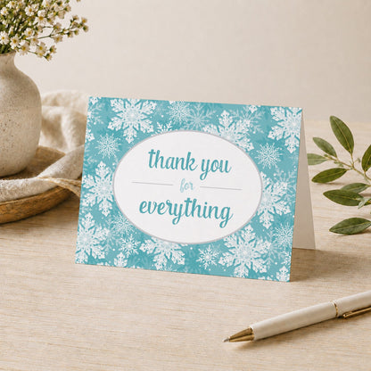 Teal Snowflake Winter Thank You Card shown on a desk with a vase, plants, and a pen.