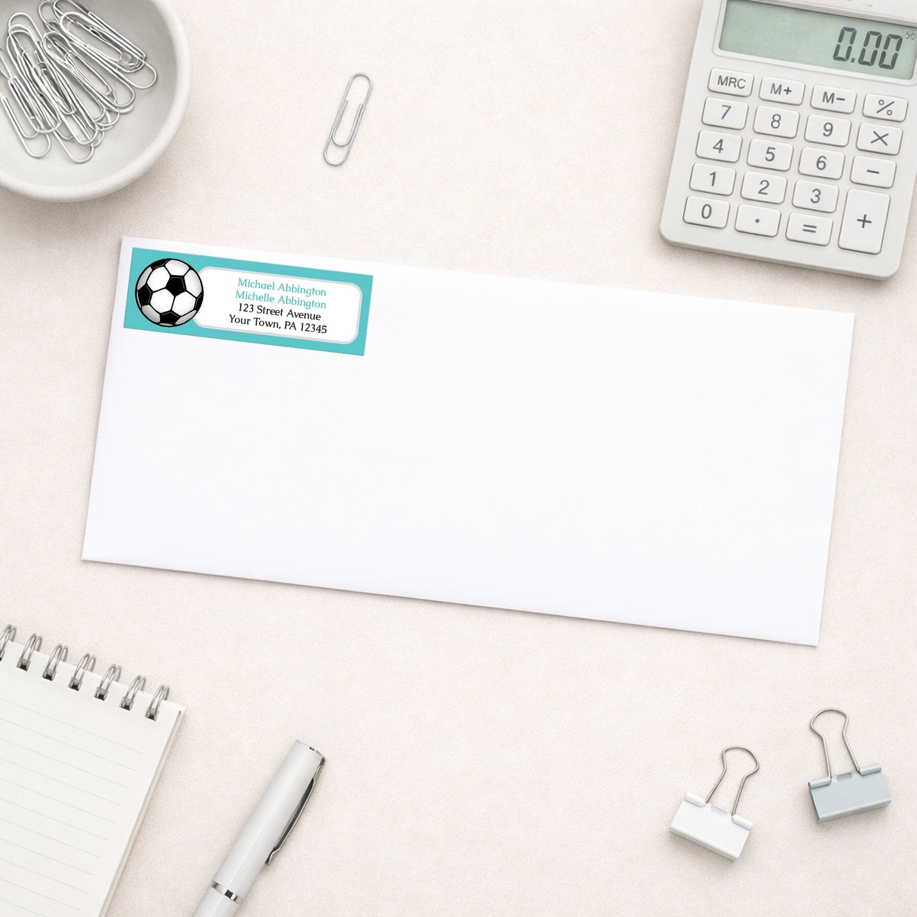 Teal Soccer Ball Return Address Label shown on a white envelope with everyday office supplies